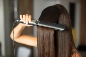 Young brunette woman straightening her glowing healthy hair using modern iron hair straightener at...
