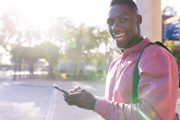 Smiling man using smartphone outdoors, wearing backpack, enjoying sunny day, copy space