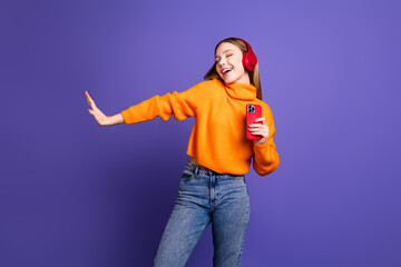 Photo of cheerful teenager girl in orange turtleneck and jeans hanging out with smartphone earphones isolated on purple color background