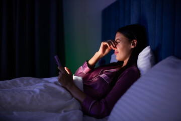 Fototapeta premium Young woman feeling eye pain while using cellphone at night, lying in bed suffering from insomnia, side view