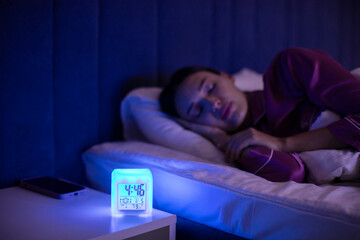 Modern digital alarm clock lamp on bedside table near sleeping young lady in morning at home, selective focus on lighting gadget