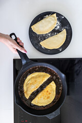 person holding a frying pan on which pies (chebureks) are fried. Near the induction cooker there is a black plate with raw pies.