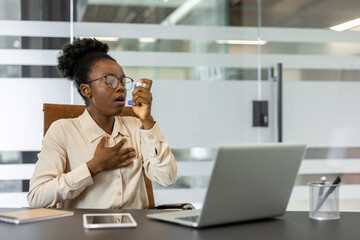 Young african american woman experiencing asthma attack in office environment uses inhaler while sitting at desk with laptop and phone nearby, demonstrating health challenges in workspaces.