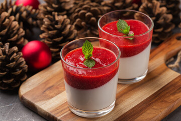 Two portion coconut Panna cotta with strawberry sauce in glasses on a dark stone table. Christmas and New Year's composition. Red Christmas balls, pine cones in background.