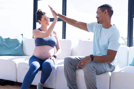 At home, multiracial couple, high-fiving, pregnant woman and man on couch, celebrating together - Powered by Adobe