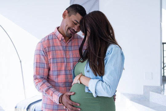At home, multiracial couple, expecting parents embracing, man touching woman's pregnant belly