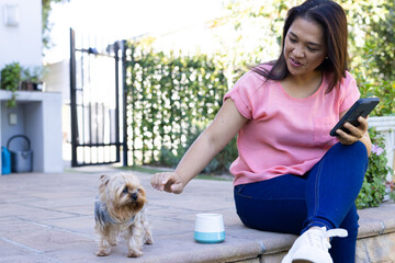 Sitting outdoors, woman using smartphone and petting small dog near automatic feeder