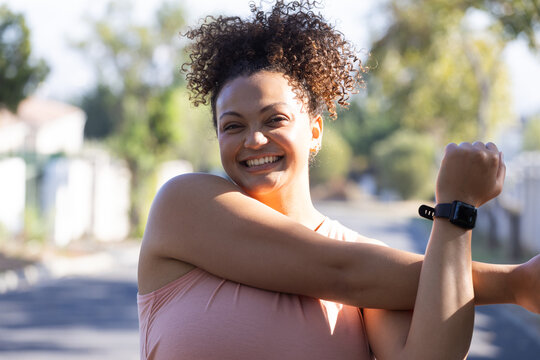 Smiling woman with curly hair stretching arms outdoors wearing smartwatch, enjoying morning exercise