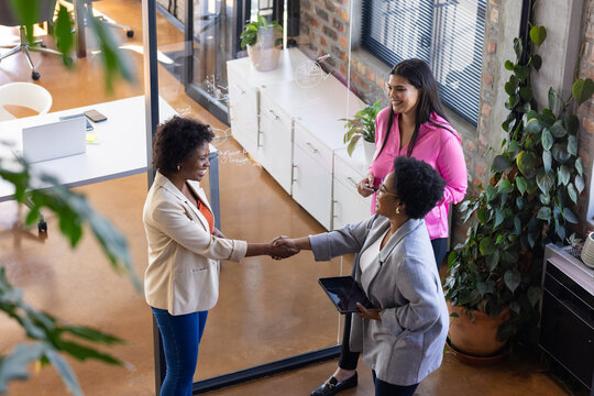 Businesswomen shaking hands in modern office, colleague holding tablet and smiling