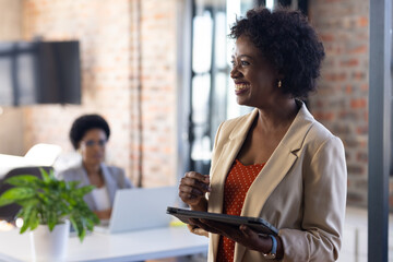 Smiling businesswoman holding tablet in modern office, colleague working on laptop
