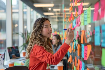 A woman in red shirt is holding up colorful sticky notes on the glass wall of an open office space.