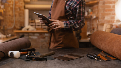 Leather craftsman uses computer tablet for communications on atelier workshop. Concept technology in DIY business banner