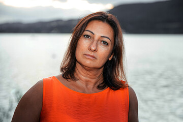 Woman with serious expression in orange dress by calm lake during cloudy sunset, peaceful outdoor...