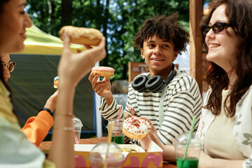 Medium shot of smiling teenage Black boy holding delicious donut sitting at cafe table with friends while enjoying spending time together outside in sunshine, copy space