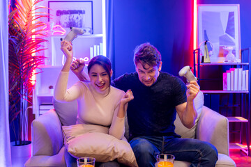 Couple joyful player video game on TV using joysticks raising fist up of winner together with snack...