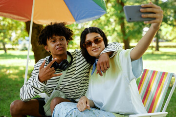 Medium shot of trendy Middle Eastern girl taking selfie with African American teenage boy while sitting in folding chairs having fun in park