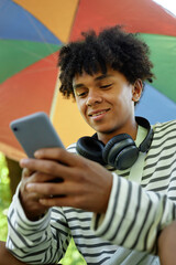 Vertical shot of smiling Black boy with headphones text messaging on phone while enjoying online communication on summer day in park