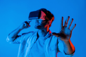 Caucasian man standing and making hand gesture and wearing VR glass. Smiling teenager using futuristic virtual reality technology goggles and display virtual program with neon background. Deviation.