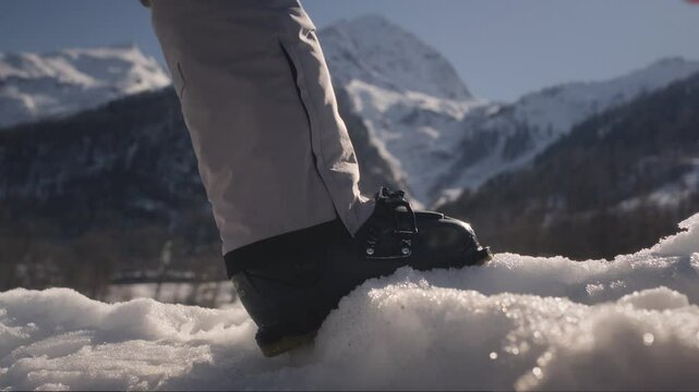 Male skier puts foot on snowdrift securing ski boots by tightening clips against snowy mountain scenery. Skilled athlete prepares equipment for upcoming skiing adventure at ski resort
