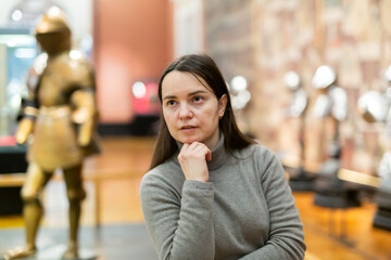 Female museum visitor examining with interest ancient iron armor displayed on exhibition ..