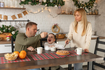 Family mom, dad and little son drinking hot chocolate, tea, coffee, cocoa in kitchen at festive table and eating home made pie. Christmas tree and home decorated interior. Happy New Year.
