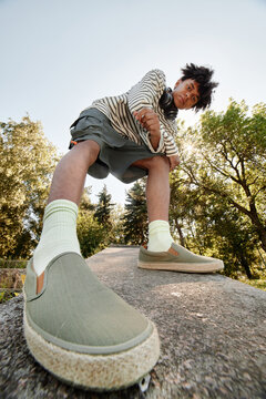 Vertical shot of cool teenage Black boy in playful mood posing for camera with foot in front while walking in city park, fisheye effect