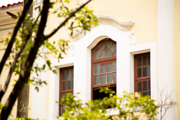 Janela, sacada, grades e paredes de casarão de arquitetura antiga cercada por galhos de árvore. 
