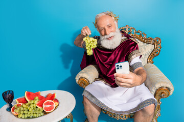 Portrait of ancient myth god smart phone make selfie hold grape sit throne isolated on blue color...