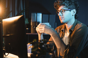 Young businessman working creative startup project job, drinking hot drink with coffee or tea while focusing on pc at neon light modern office. Thinking marketing plan online at night time. Gusher.
