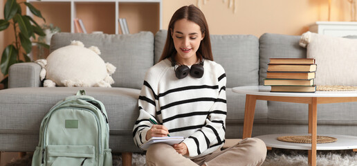Beautiful happy female student with stack of books, backpack and headphones studying on floor at...