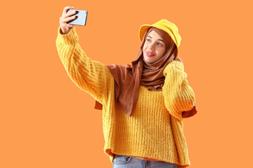 Young Muslim woman with mobile phone taking selfie on orange background