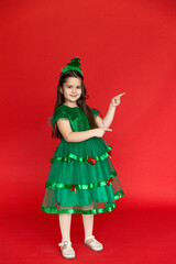 A girl in a festive carnival costume of little Christmas tree pointing at an empty space on a red background. Winter holidays concept. Sale and advertising