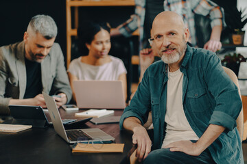 A man in a blue shirt sits at a table with a laptop and a few other people. He is smiling and he is enjoying the company of his colleagues