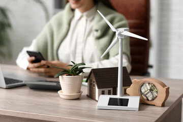 Wind turbine with house model, piggy bank and plant on table of female engineer in office, closeup