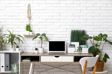 Workplace with blank laptop and green plants near white brick wall in office