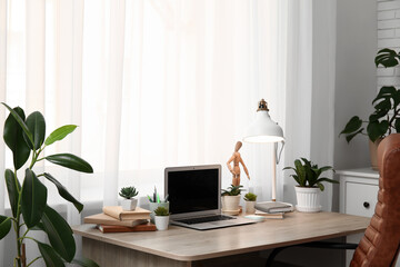 Workplace with blank laptop, lamp and green plants near window in office