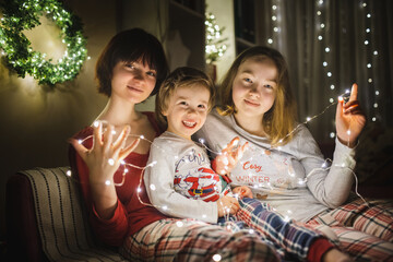 Two big sisters and their toddler brother playing with Christmas lights in a cozy living room on Christmas eve. Kids spending time at home during winter break.