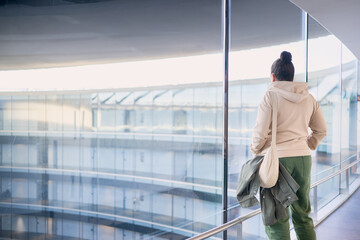 young woman, seen from behind, gazes thoughtfully out of a large floor-to-ceiling window in a modern hospital. Contemporary architecture