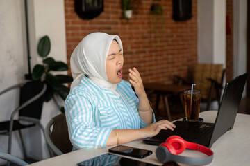 a young Asian female worker wearing a hijab who looks exhausted while working at the cafe, the concept of remote working, working flexibly from anywhere.
