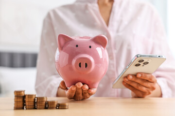 Woman with piggy bank and smartphone at table indoors, closeup