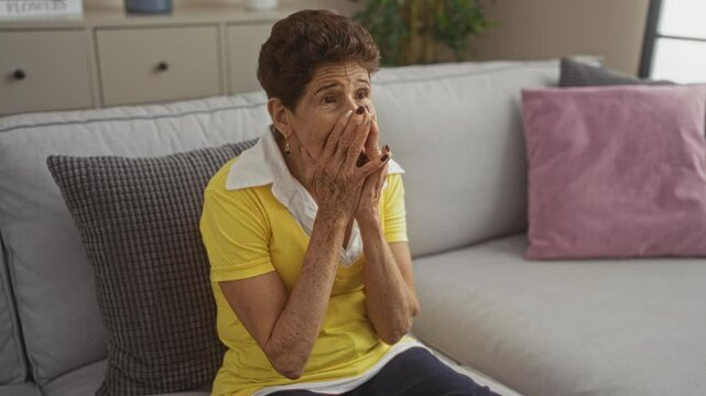 A mature, hispanic woman wearing a yellow shirt looks distraught while sitting on a sofa in her living room.