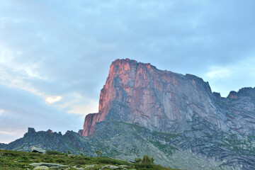 A high rock with a steep steep slope is painted orange by the setting sun on a warm summer evening.