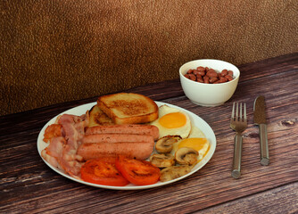 A plate with scrambled eggs, croutons, sausages and fried bacon, mushrooms, tomatoes and beans on a wooden table, with cutlery nearby.