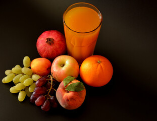 A mixture of fruit juices in a tall faceted glass on a black background, next to ripe fruits, apple, orange, peach, pomegranate and bunches of grapes.