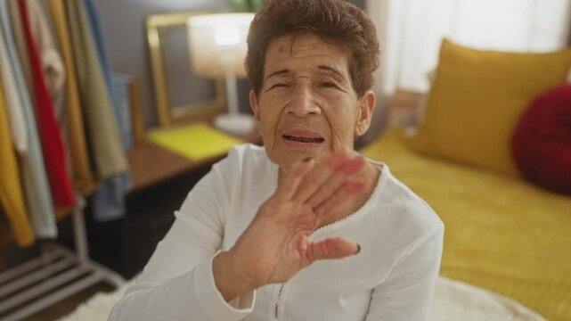 Elderly hispanic woman with short hair in a white blouse sends a kiss and waves at the camera, seated in a cozy room with a bed and clothes hanging in the background.