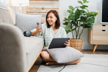 Fototapeta premium Smiling young woman sitting on the floor in a cozy living room, using a smartphone and tablet, surrounded by modern decor.