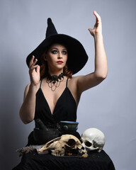 Close up portrait of beautiful  female model wearing black dress, pointy witch hat halloween costume.  Isolated figure on studio background. Casting magic spell at spooky occult  ritual potion table 