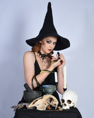 Close up portrait of beautiful  female model wearing black dress, pointy witch hat halloween costume.  Isolated figure on studio background. Holding snake serpent at spooky occult ritual potion table 