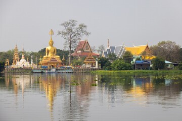 A serene landscape featuring a large golden Buddha statue beside traditional Thai temples, reflected in calm waters. Lush greenery surrounds the area, creating a peaceful atmosphere.