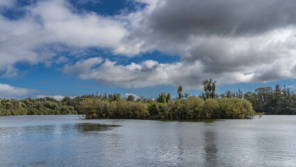 Obraz premium In the middle of a calm lake, an island covered with green vegetation is visible. Ripples on the water. Clouds in the blue sky. Mauritius. Grand Bassin. Ganga Talao